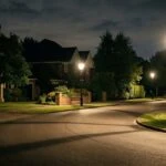 Wide context view of a quiet, wealthy suburban street at night with glowing streetlamps, manicured lawns, and deep