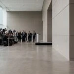 Wide shot of a spacious, modern hall with large windows. A group of media personnel with cameras and tripods stands